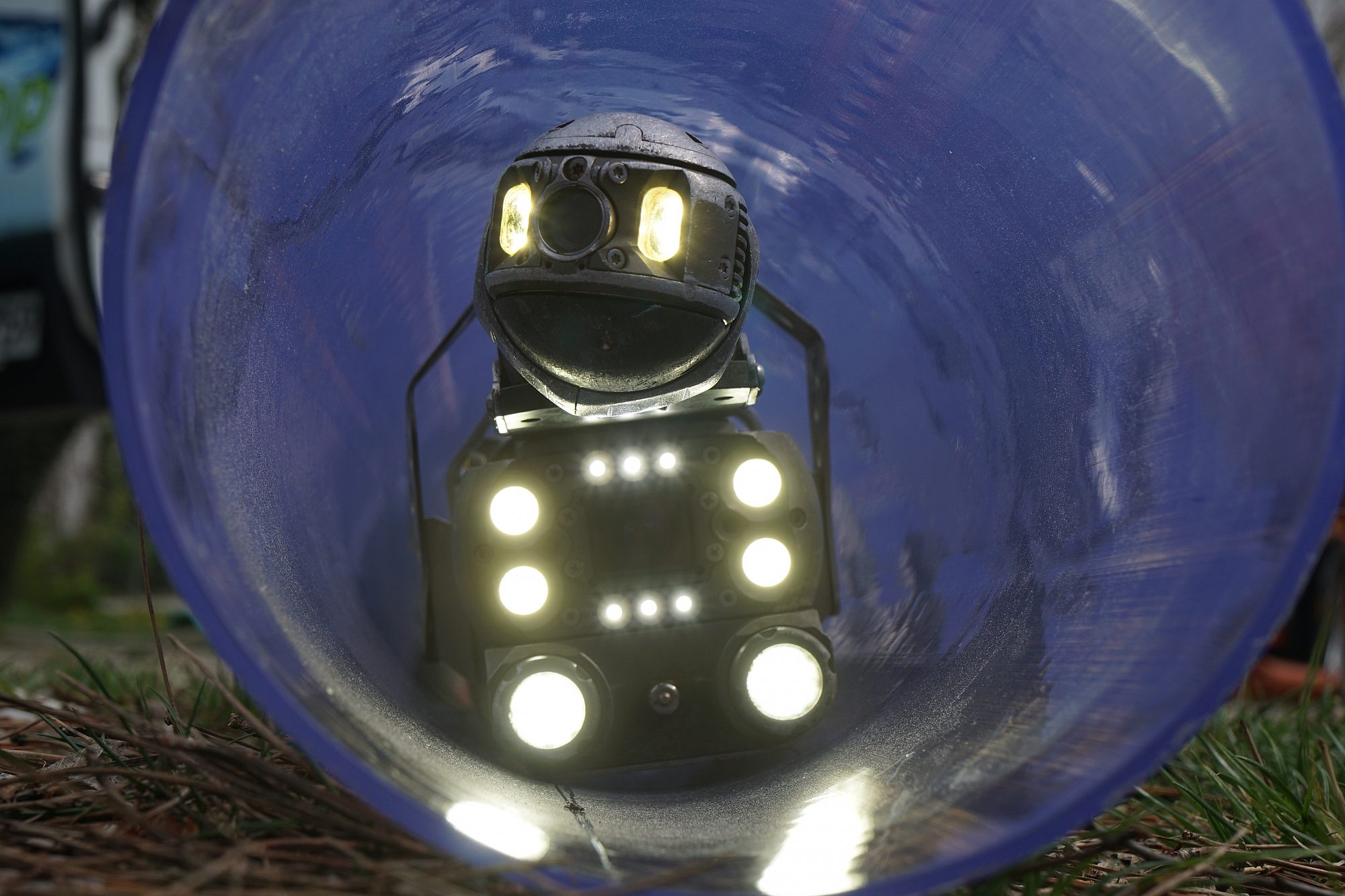 Der Einblick in ein Rohr zeigt einen Kanalroboter mit vielen Lampen, der gerade auf den Betrachter zufährt.
